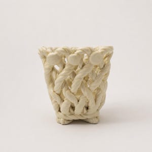 May include: A cream-colored ceramic vessel featuring an intricate, open-work lattice design that mimics the appearance of braided rope. The structure is cylindrical with a flared top rim and rests on a base formed by the woven ceramic strands.