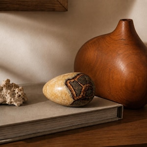 May include: A smooth, egg-shaped stone with a polished tan and dark grey veined surface, positioned next to a piece of porous white coral and a large, rounded wooden vase with a smooth, warm brown finish on a closed, light grey hardcover book.