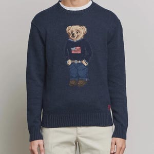 May include: A navy blue long-sleeve crewneck sweater featuring a knitted graphic of a teddy bear wearing a dark blue sweater with a small American flag design, blue jeans, and brown shoes, with red embroidered letters near the bottom hem.