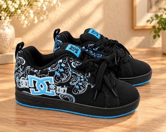 May include: Black low-top sneakers featuring a light blue DC logo on the side, black laces, and a patterned design with white and light blue swirls. The shoes have a black sole with a light blue trim and a black tongue with a blue logo patch.