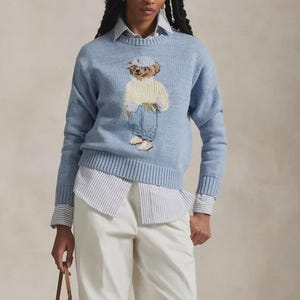 May include: A light blue long-sleeve knit sweater featuring a central graphic of a teddy bear wearing a white cap, a cream-colored cable knit sweater, and blue jeans. The sweater is layered over a white and blue striped button-down shirt.