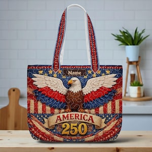 May include: A patriotic tote bag featuring a bald eagle with outstretched wings, set against a red, white, and blue background. The bag has a banner that reads "AMERICA 250". The handles are red and blue with white stitching. The bag has a small tag that says "Name".