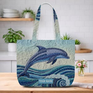 May include: A blue and white quilted tote bag featuring a leaping dolphin over ocean waves. The bag has a light blue background with a dolphin design in shades of blue. The text "YOUR NAME" is displayed on the bottom of the bag.
