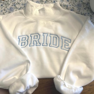 May include: A white long-sleeve sweatshirt featuring a mock neck collar and the word BRIDE embroidered across the chest in light blue block lettering with a white outline. The garment has long sleeves with ribbed cuffs and a ribbed hem.