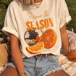 Puede incluir: Camiseta color crema con la frase naranja "Survive the SEASON", una bola de discoteca y rodajas de naranja. La camiseta se combina con pantalones cortos vaqueros. También se ve una bebida en un tarro de cristal.