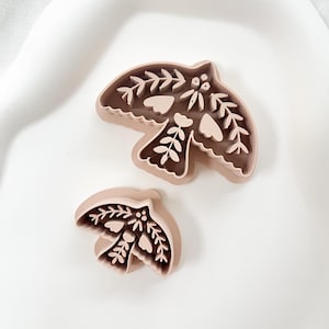 May include: Two bird-shaped cookie cutters. The cutters are light brown and feature detailed floral and leaf designs. The larger cutter measures approximately 10 cm wide, while the smaller one is about 6 cm wide. They are placed on a white surface.