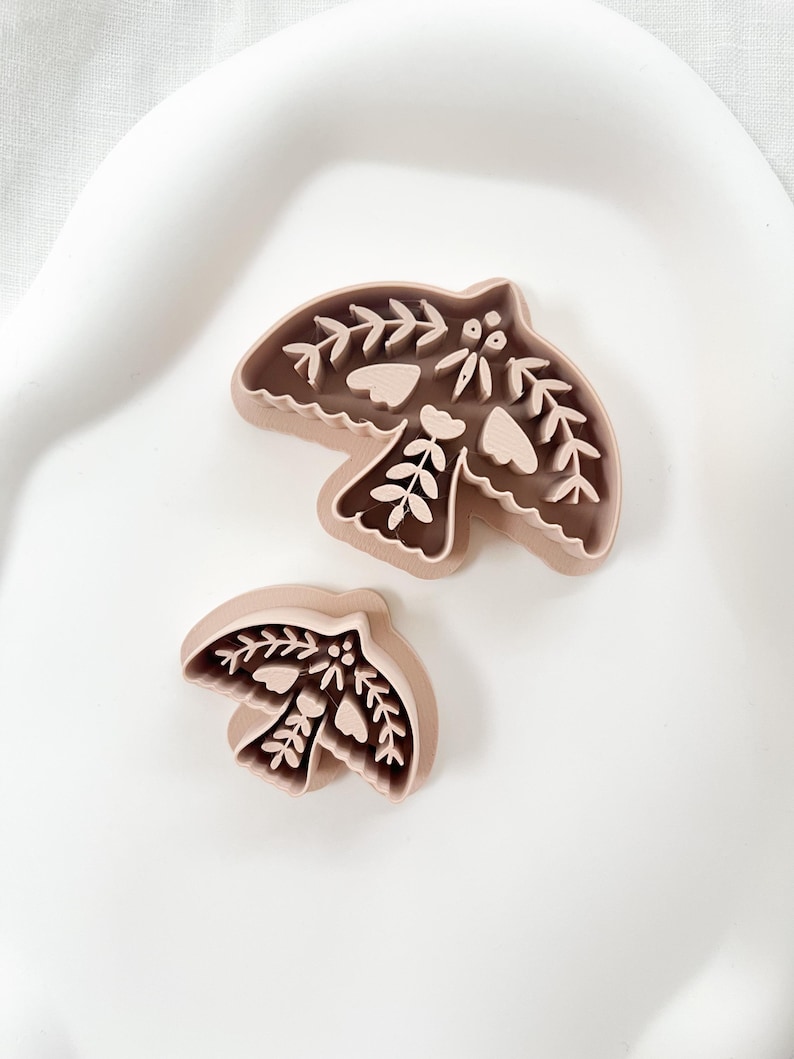 May include: Two bird-shaped cookie cutters. The cutters are light brown and feature detailed floral and leaf designs. The larger cutter measures approximately 10 cm wide, while the smaller one is about 6 cm wide. They are placed on a white surface.