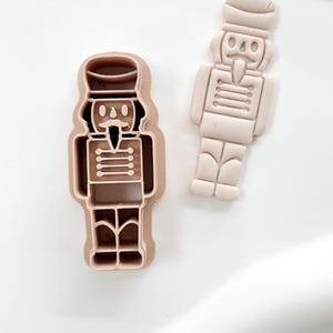 Puede incluir: Dos cortadores de galletas de plástico marrón claro con forma de soldado cascanueces. Uno es un contorno sólido y el otro es una impresión detallada del mismo diseño. Los cortadores están sobre una superficie blanca.