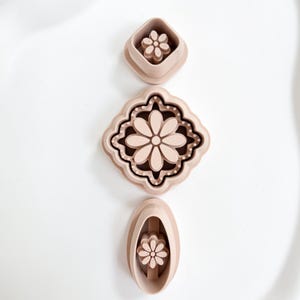 Könnte beinhalten: Drei beige Ausstecher in Blumenform in verschiedenen Designs. Der obere Ausstecher ist quadratisch, der mittlere hat eine dekorative Form und der untere ist oval. Die Ausstecher sind vertikal auf einer weißen Oberfläche angeordnet.