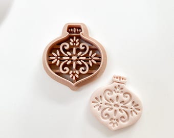 May include: Two light brown Christmas ornament-shaped cookie cutters. One cutter is a frame with a detailed floral design, and the other is a solid ornament with the same design. The cutters are on a white surface.