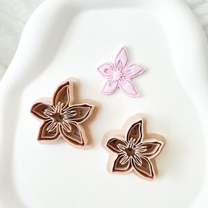 以下が含まれることがあります： 3つの花型のクッキーカッター。2つは茶色の濃淡で重ねられ、1つは無地のピンク色です。カッターは白い曲面の上に配置されており、ベーキングやクラフトをテーマにしていることを示唆しています。