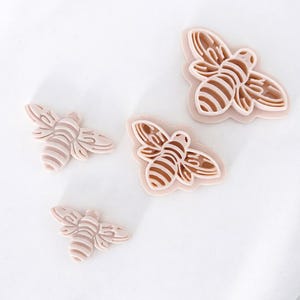 May include: Four light brown bee-shaped cookie cutters. The cutters are in various sizes and have detailed wing and body designs. The bee cutters are on a white surface.