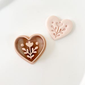 Op de afbeelding: Twee hartvormige koekjessnijders. Eén is lichtbruin met een bloemenontwerp in het midden. De andere is lichtroze met hetzelfde bloemenontwerp. De uitstekers liggen op een wit oppervlak.