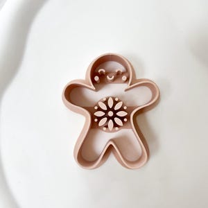 Pode incluir: Cortador de biscoitos em forma de homem de gengibre castanho claro com um rosto sorridente e um desenho floral no centro. O cortador é feito de plástico e está sobre uma superfície branca. Projetado para fazer biscoitos.
