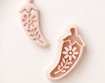 May include: Two pale pink chili pepper shaped cookie cutters. One cutter is a solid stamp with a debossed floral pattern featuring a central flower and leaves. The second cutter is an open frame outline that matches the shape and design of the stamp.