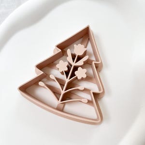 May include: A light brown, tree-shaped cookie cutter with a floral design. The cutter features a detailed outline of a fir tree with a sprig of flowers and leaves in the center. The cutter is made of plastic and sits on a white surface.