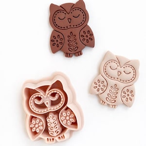 May include: Three owl-shaped cookie cutters and a finished biscuit. The cookie cutters are light pink, and the finished biscuit is light brown. The owl design includes closed eyes, floral and leaf patterns, and a detailed outline.