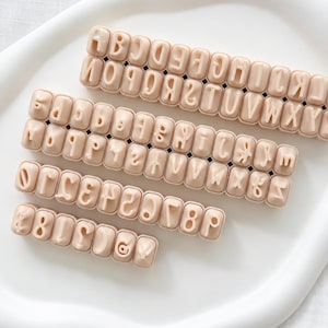 May include: A set of light beige plastic alphabet and number stamps arranged in four rows. The top row features uppercase letters A through Z. The second and third rows contain lowercase letters, while the bottom row displays numbers and symbols.