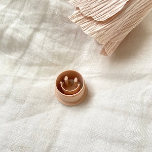 Könnte beinhalten: Ein hellbrauner Plastik-Ausstecher in Form eines Smiley-Gesichts mit zwei kleinen Löchern für die Augen und einer geschwungenen Linie für den Mund.