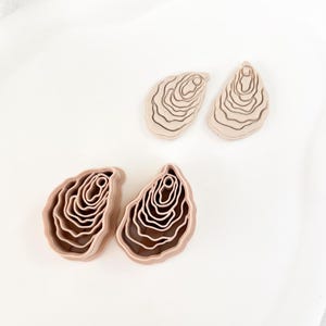 Può includere: Quattro taglia biscotti in argilla polimerica beige a forma di ostrica. Ogni taglia biscotti presenta un design a strati e scanalato, che ricorda l'interno di un guscio di ostrica. I taglia biscotti sono disposti su una superficie bianca.