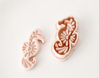May include: Two pale pink seahorse-shaped tools, one flat stamp and one deeper cutter, both featuring an embossed floral and leaf pattern on their surfaces. The items are light pink in color and display a detailed, stylized seahorse silhouette.