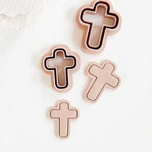 Puede incluir: Un conjunto de cuatro cortadores de galletas en forma de cruz de diferentes tamaños. Los cortadores son de color rosa claro con un contorno rosa más oscuro. Un cortador tiene un contorno negro. Están dispuestos sobre una superficie blanca.