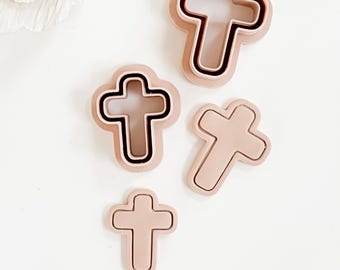 May include: A collection of four cross-shaped cookie cutters in varying sizes. The cutters are a light pink color with a darker pink outline. One cutter has a black outline. The cutters are arranged on a white surface.