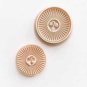 Può includere: Due timbri circolari in argilla color beige chiaro con un design a raggiera solare e un volto umano stilizzato al centro. Il timbro più grande è posizionato sopra quello più piccolo, entrambi mostrano linee radianti dal centro.