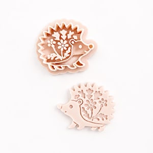 Peut inclure: Deux emporte-pièces en plastique rose clair en forme de hérisson. Chaque pièce présente un motif en relief sur le dessus représentant un hérisson avec des fleurs et des feuilles sur le flanc, l'un tourné vers la droite et l'autre vers la gauche.