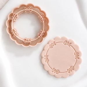 May include: A light peach-colored circular cookie cutter with a scalloped edge and an internal leaf wreath design, accompanied by a matching circular cookie stamped with the same leaf wreath pattern, both featuring a smooth, matte finish.