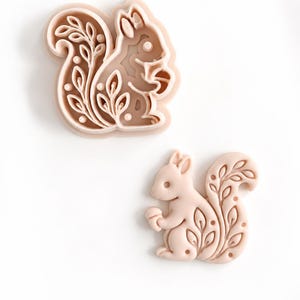 May include: A light peach-colored squirrel-shaped cookie cutter and a matching embossed squirrel-shaped cookie stamp. Both items feature a detailed design of a squirrel holding an acorn with intricate leaf patterns on its tail.