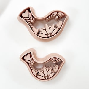 May include: Two light pink bird-shaped cookie cutters. Each cutter features a detailed interior design with hearts, leaves, and dots. The cutters are made of a sturdy material and are ideal for baking themed cookies.