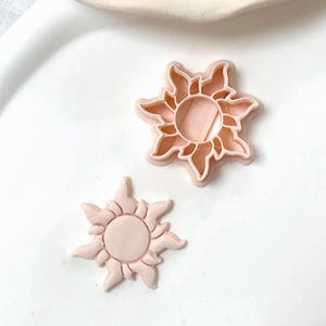 Peut inclure: Un emporte-pièce en forme de soleil de couleur pêche clair et une pièce découpée assortie en forme de soleil. L'emporte-pièce présente un motif circulaire central entouré de huit rayons stylisés en forme de flammes, reproduits sur la pièce.