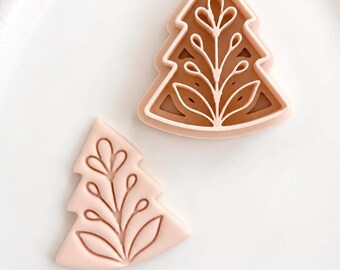 May include: Two light pink cookie cutters shaped like Christmas trees. The larger cutter has a brown interior insert, while the smaller one is solid light pink. Both feature a raised, intricate botanical leaf pattern on their surfaces.