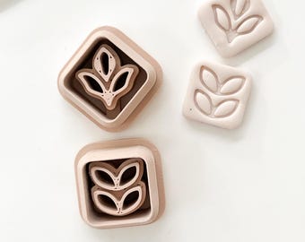 May include: A set of four light brown leaf-shaped cookie cutters. Two cutters are square, and two are diamond-shaped. The leaf design is embossed on the square cutters and is a three-dimensional design on the diamond-shaped cutters.