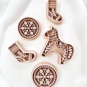 May include: A collection of five light pink cookie cutters in various holiday shapes. The shapes include two snowflake designs, two Christmas stockings, and a horse. The cookie cutters are arranged on a white surface.