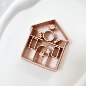 May include: A light brown house-shaped cookie cutter with a bird, floral, and circle design. The cutter has rectangular windows, a door with a flower, and a roof. The cutter is made of plastic.