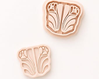 May include: Two light peach-colored cookie cutters with a symmetrical floral design. Each cutter features a central stem flanked by two stylized flower buds and curved leaves, creating a cohesive botanical pattern in a flat, molded plastic form.