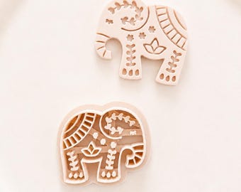 May include: Two light beige elephant-shaped cookie cutters. Each cutter features intricate embossed patterns including floral motifs, stripes, and geometric shapes on the body, trunk, and legs, designed to imprint details into dough.
