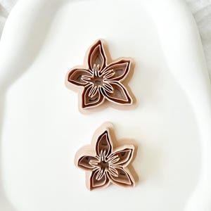 以下が含まれることがあります： 薄茶色の花型のクッキーカッターが2つ。カッターは、各層が前の層よりわずかに小さく、3次元効果を生み出す、層状のデザインです。カッターは白い表面にあります。