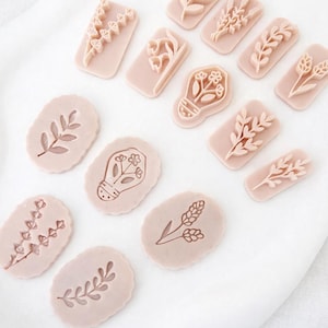 May include: A collection of pink floral-themed stamps in various shapes. The stamps feature detailed designs of leaves, flowers, and sprigs. The stamps are arranged on a white surface, showcasing their intricate patterns.