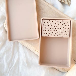Puede incluir: Una caja de almacenamiento rectangular de color rosa claro con una bandeja extraíble. La bandeja tiene una cuadrícula de pequeños agujeros.