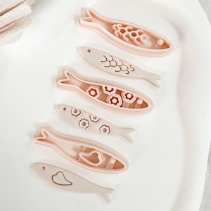 May include: Six fish-shaped cookie cutters in varying designs and colours. The cutters are light pink and beige, with decorative interior cutouts. The fish shapes include designs such as bubbles, flowers, and hearts. The cutters are arranged on a white oval tray.