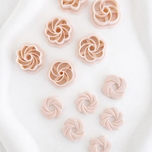 Peut inclure: Diverses découpoirs à biscuits en forme de fleurs rose clair, disposés sur une surface blanche. Les découpoirs présentent différents motifs, tels que des spirales et des pétales. Idéal pour la pâtisserie et l'artisanat.