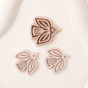 Könnte beinhalten: Drei vogelförmige Ausstechformen oder Stempel in hellem Beige. Jedes Teil zeigt ein stilisiertes Vogeldesign mit linearen Flügel- und Schwanzdetails. Die Formen sind so angeordnet, dass sich ein größeres Teil oben und zwei kleinere Teile darunter befinden.