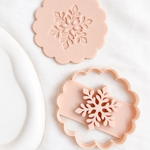 May include: A light peach-coloured cookie cutter set featuring a scalloped circular frame and a separate insert with a detailed snowflake design. The set includes one stamped impression piece and one outer cutting frame with a snowflake centre.