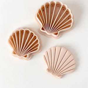 Pode incluir: Três cortadores de biscoitos em forma de concha na cor pêssego claro. Dois dos cortadores têm um design de estrutura aberta com nervuras internas, enquanto o terceiro é um carimbo de face sólida com linhas radiais em relevo.