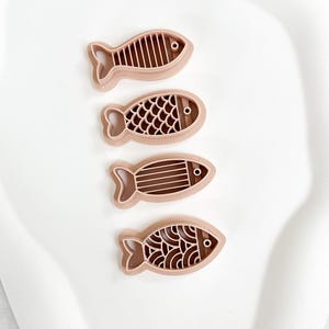 Puede incluir: Cuatro cortadores de galletas con forma de pez marrón con diferentes diseños internos. Cada cortador tiene una cola y un detalle de ojo. Los cortadores están dispuestos sobre una superficie blanca.