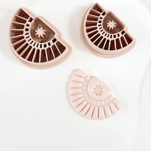 May include: Three light pink, fan-shaped cookie cutters. Two cutters have a star-shaped center and a series of vertical lines. The third cutter is a smaller, solid pink fan shape with a star in the center.