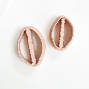 Può includere: Due formine per biscotti rosa chiaro, di forma ovale, con un centro verticale decorativo. Le formine sono in plastica e poggiano su una superficie bianca. Il design è a forma di foglia o petalo, ideale per la cottura.